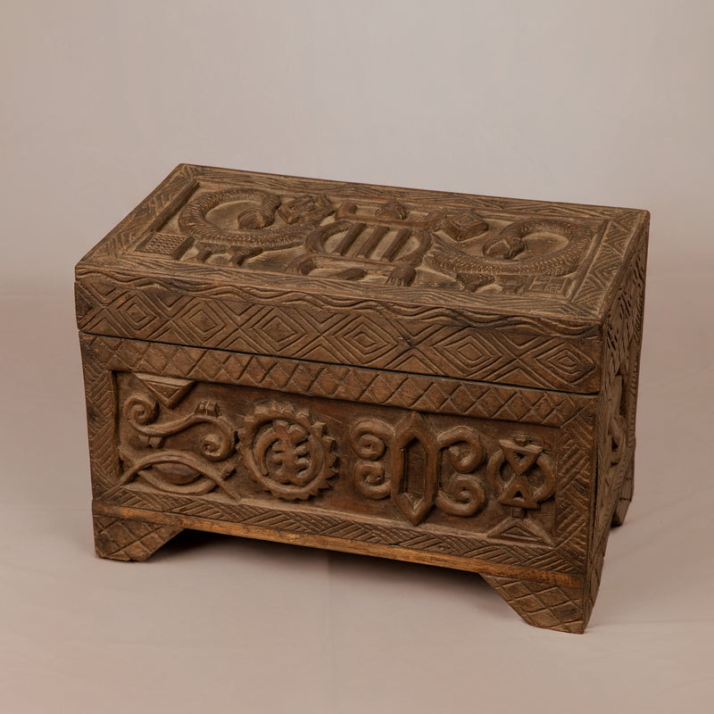 An antique, rectangular Ashanti wooden treasure box made of dark natural hardwood with a rich patina. The box has a flat, hinged lid and is completely covered in hand-carved, geometric Adinkra symbols on the top cover and all four sides. The wood shows a deep, aged luster with soft highlights on the raised carvings and darker shading in the recessed grooves. It sits on a flat surface, photographed from a three-quarter angle to show the top and two sides.